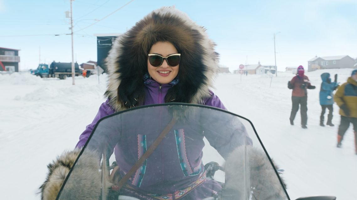 "Woman in a fur-lined hooded parka and sunglasses rides a snowmobile in a snowy village; people in winter clothing and houses are visible in the background under a bright sky."
