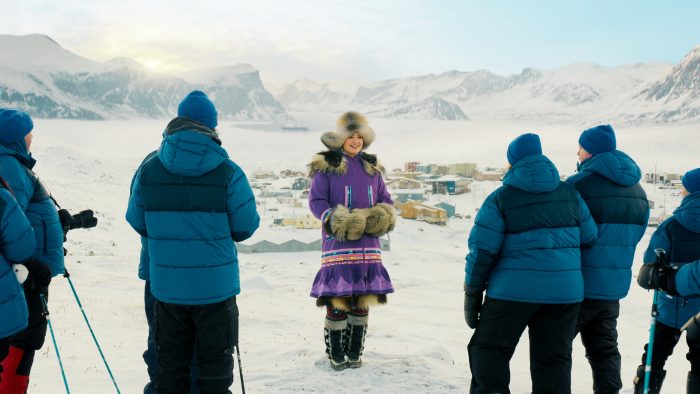 "A woman in traditional fur-trimmed clothing stands smiling in snowy arctic landscape, surrounded by five people in blue winter jackets, with a mountainous village in the background under a bright sky."